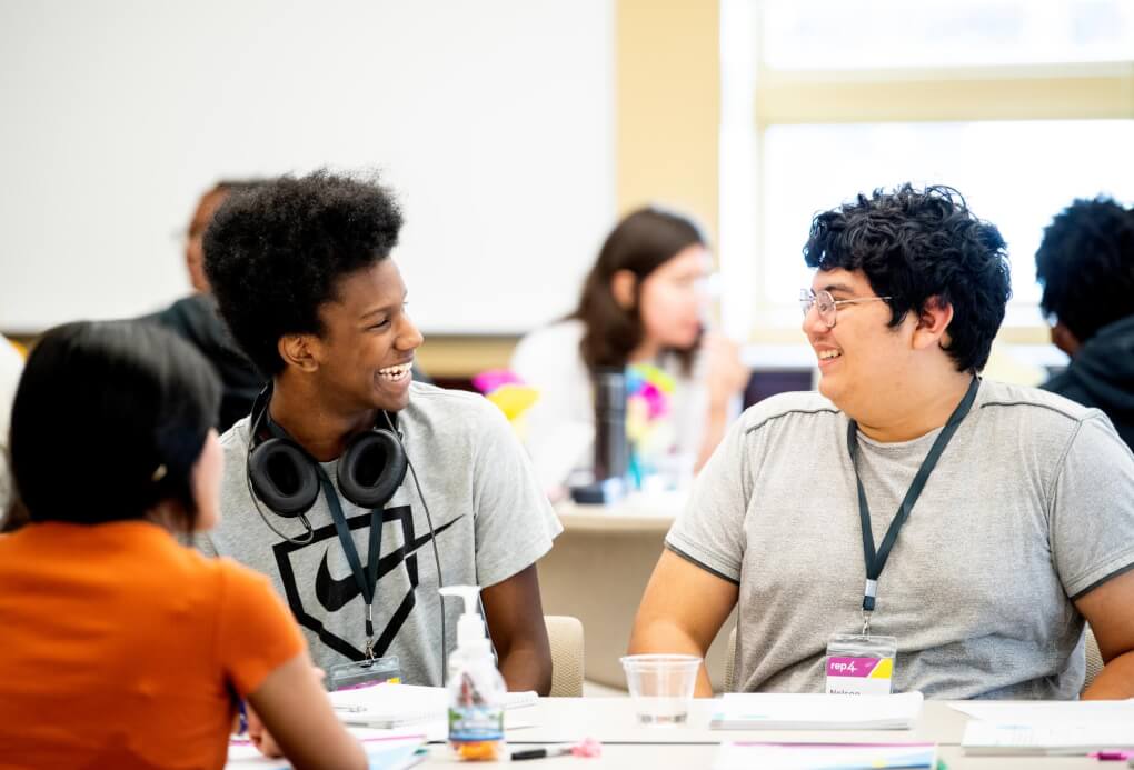 Students look at each other and laugh, in the background a classroom environment